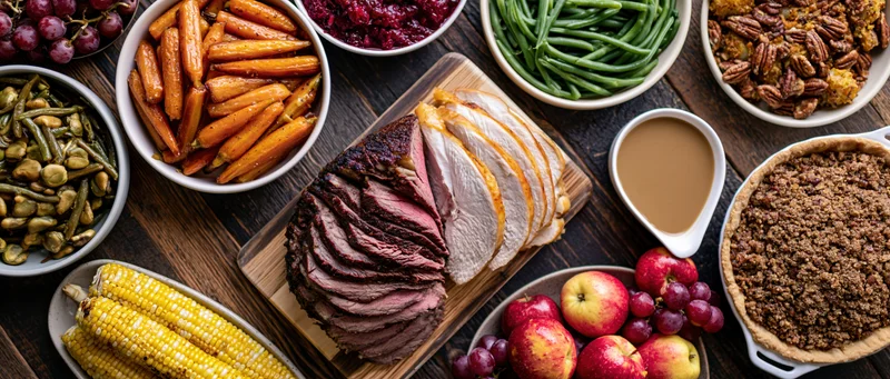 Sliced turkey and roast beef on a wooden board surrounded by holiday side dishes and desserts on a dark wooden table