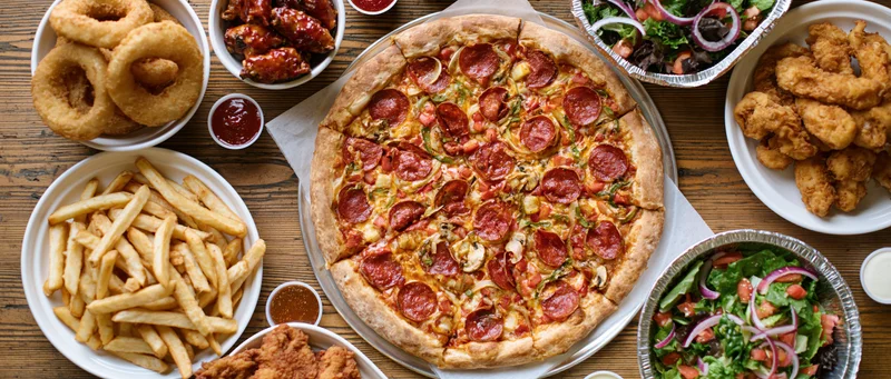 Large pepperoni and vegetable pizza with side dishes arranged on wooden table in overhead flat lay view