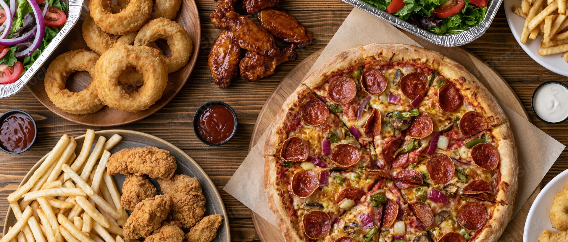Large pepperoni and vegetable pizza on wooden table surrounded by fries, onion rings, chicken wings, and dipping sauces