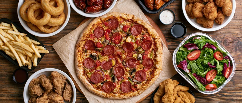 Large pepperoni and vegetable pizza surrounded by various fast food sides on wooden table