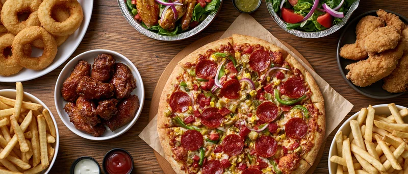 Overhead flat lay of a large pepperoni and vegetable pizza on a wooden table surrounded by side dishes including fries, onion rings, chicken wings, fried chicken, salads, and condiments