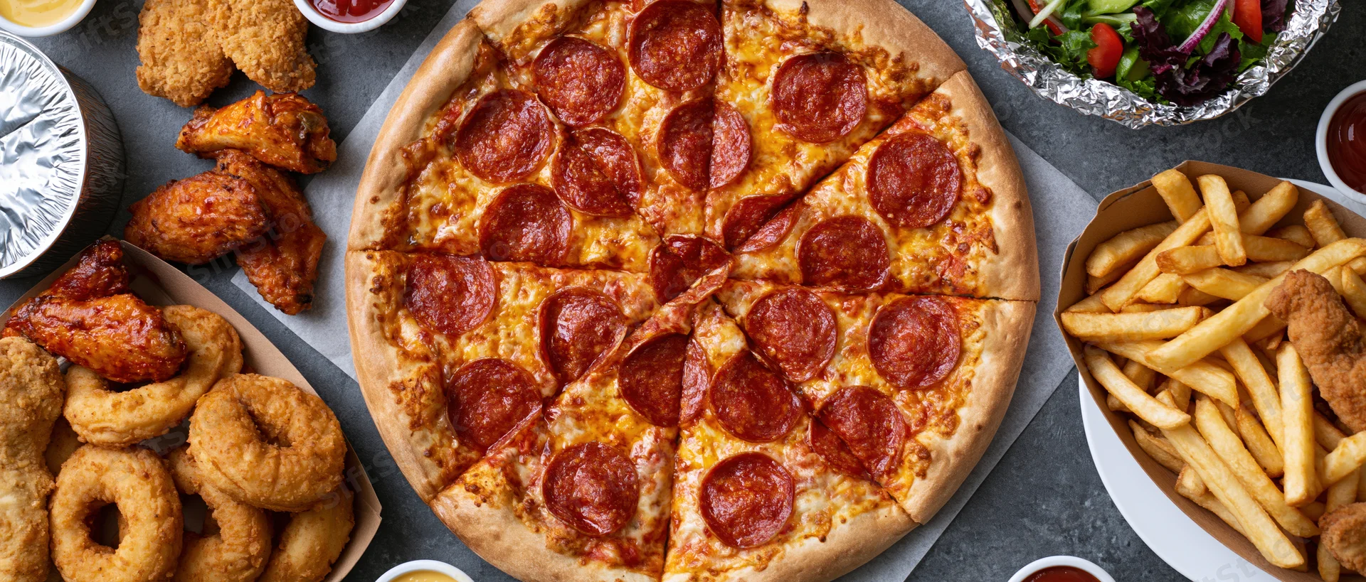 Overhead view of a large sliced pepperoni pizza surrounded by fried chicken wings, french fries, onion rings, and a salad on a gray surface