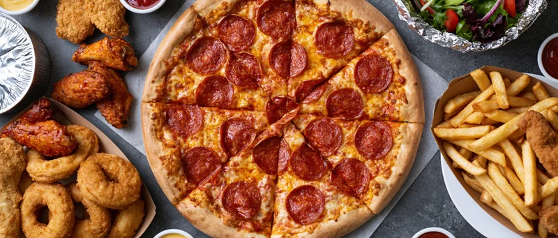 Overhead view of a large sliced pepperoni pizza surrounded by fried chicken wings, french fries, onion rings, and a salad on a gray surface
