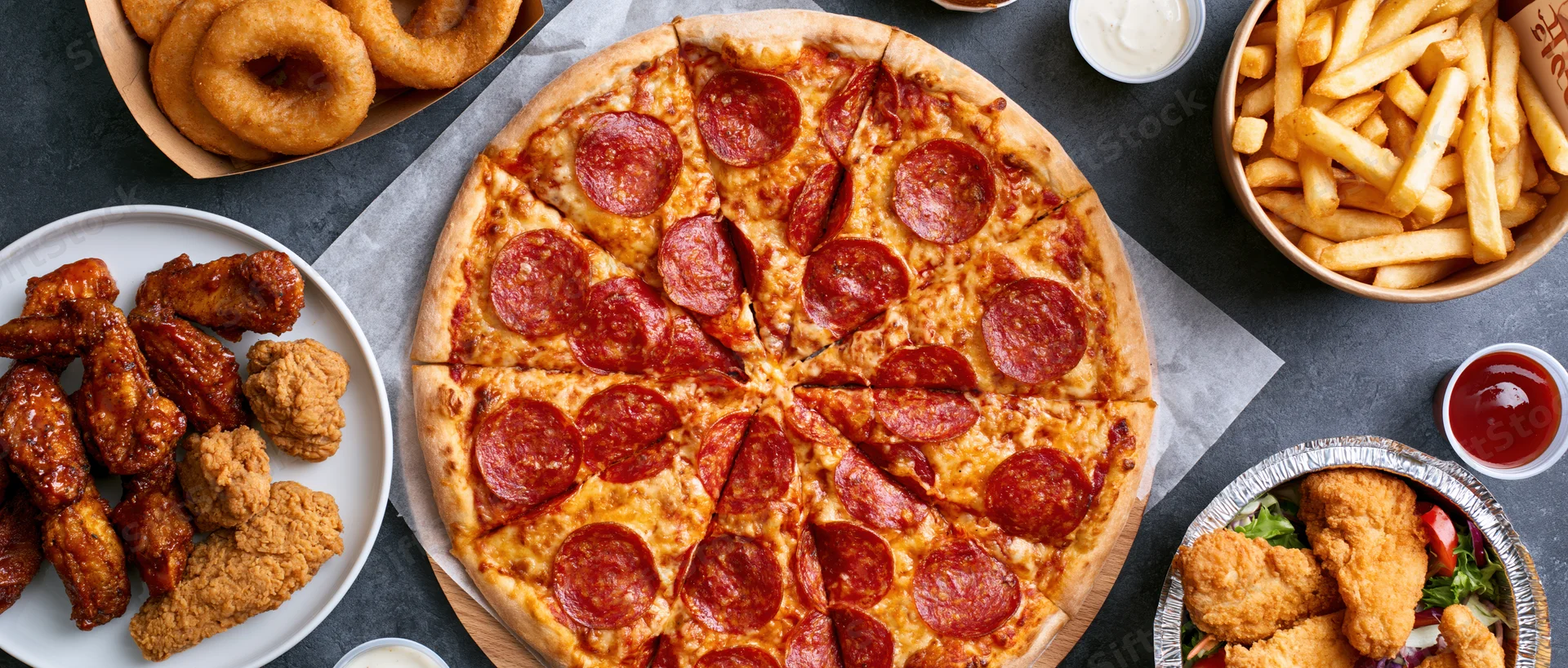 Large pepperoni pizza sliced and arranged on parchment paper with chicken wings, fries, and dipping sauces on dark table