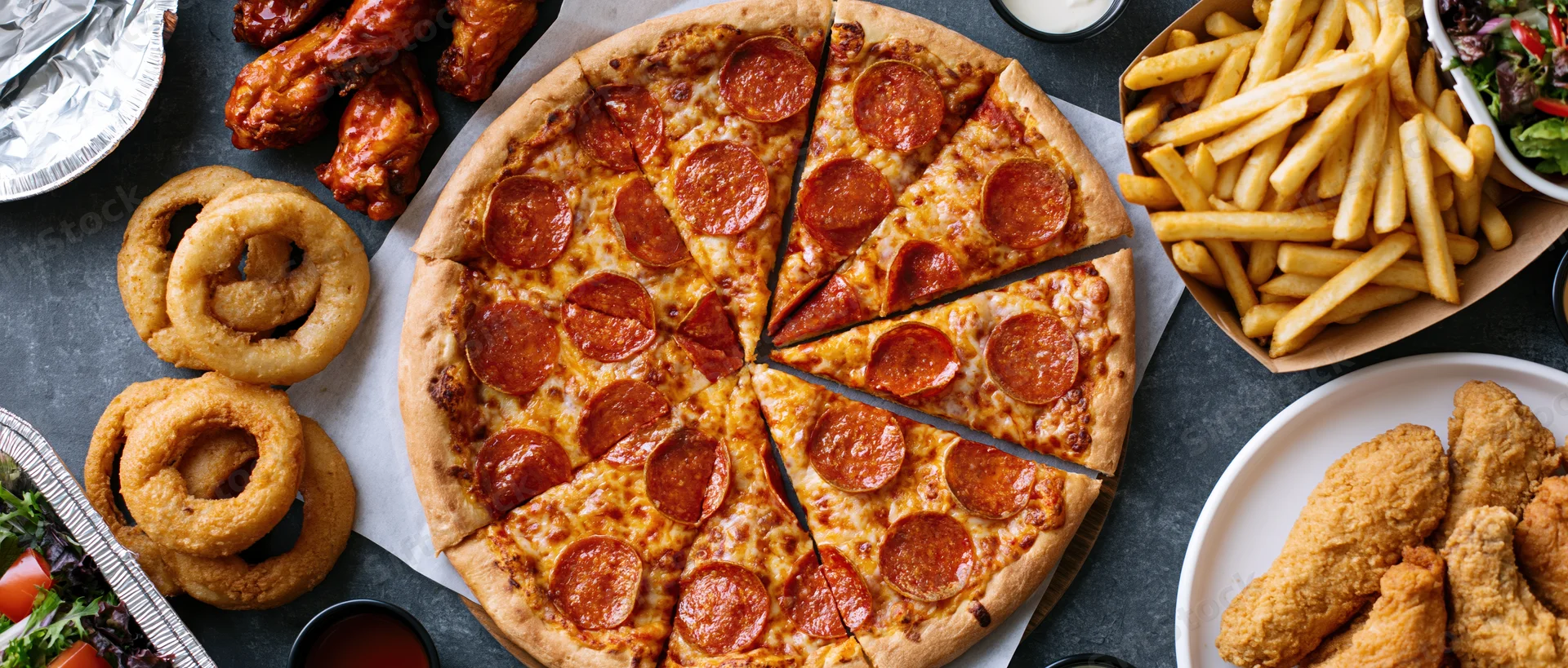 Overhead flat lay of a large sliced pepperoni pizza with fries, onion rings, chicken wings, and salad on a dark table