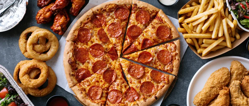 Overhead flat lay of a large sliced pepperoni pizza with fries, onion rings, chicken wings, and salad on a dark table