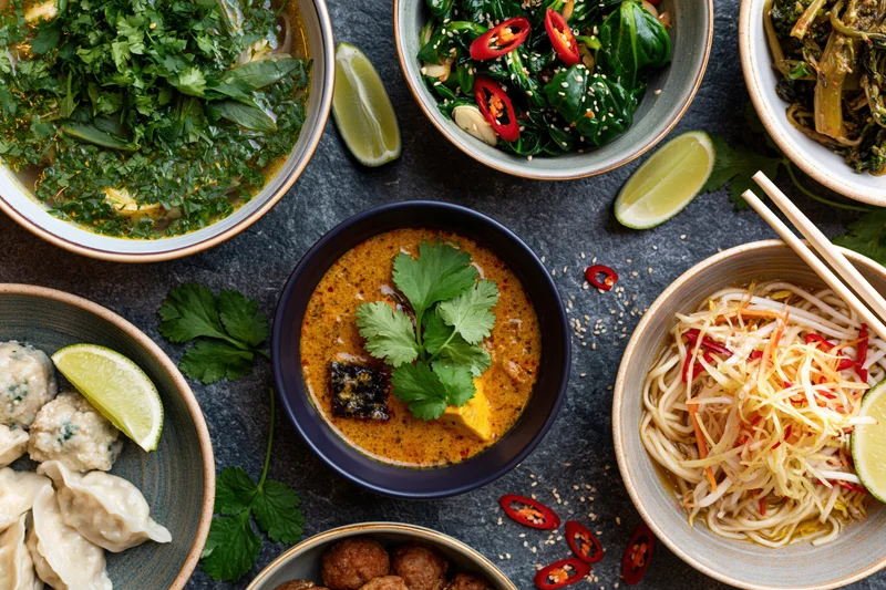 Overhead flat lay of southeast asian dishes arranged on dark stone surface with bowls of curry, noodles, dumplings, vegetables, and garnishes