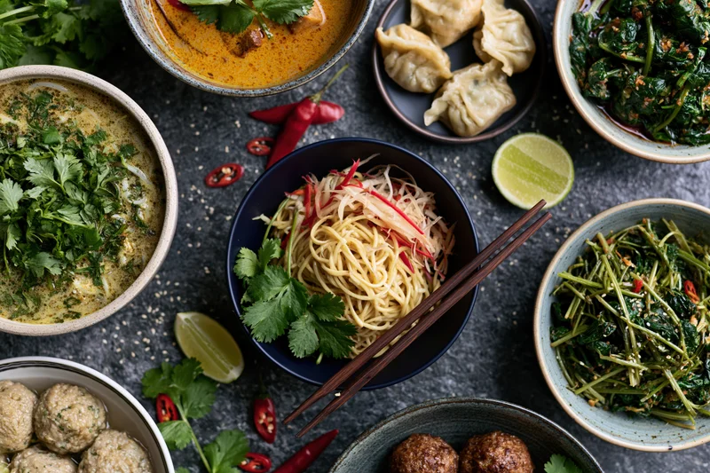Overhead flat lay of southeast asian dishes arranged on dark stone surface with bowls of noodles, dumplings, meatballs, vegetables, herbs, lime, chili, and chopsticks