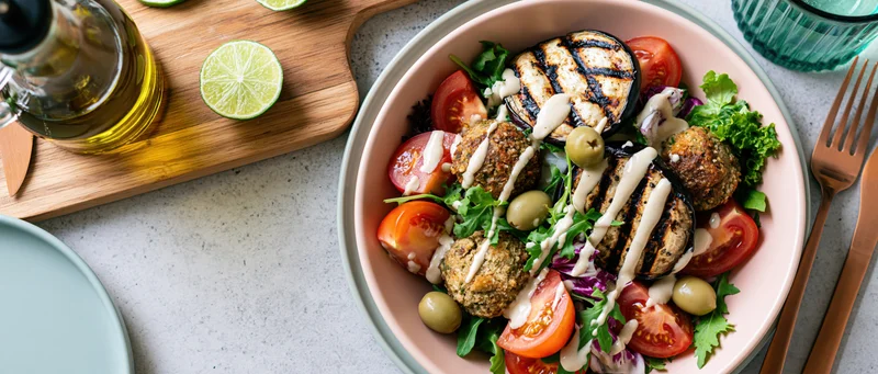 Pink ceramic bowl filled with falafel salad topped with grilled eggplant, tomatoes, olives, and dressing on a countertop