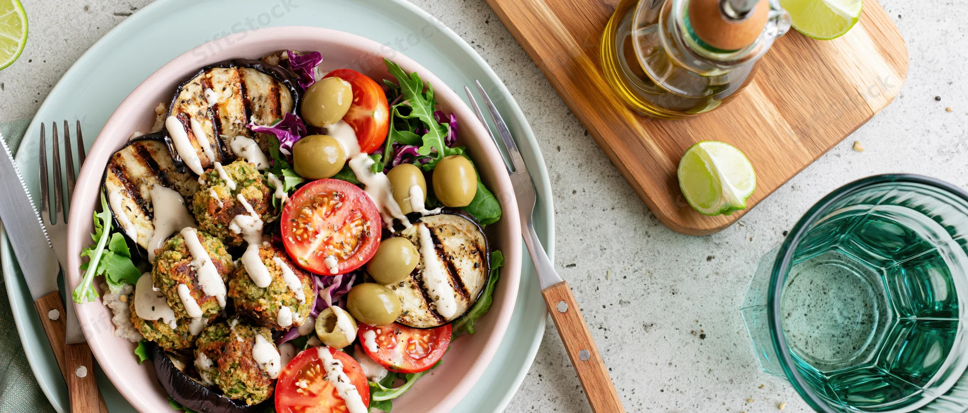 Overhead view of a pink ceramic bowl filled with falafel, grilled eggplant, tomatoes, olives and greens on a countertop with utensils and a glass of water nearby