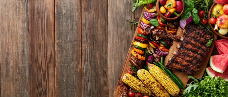 Grilled ribs and vegetables arranged on a wooden board with fruit and corn for a summer barbecue spread