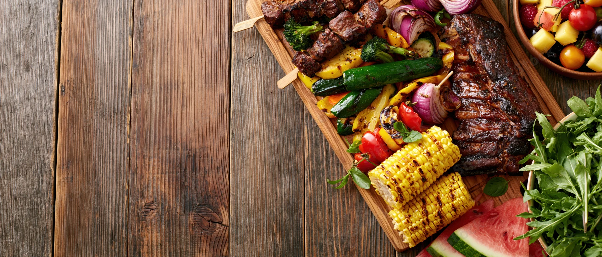 Grilled meat and vegetables arranged on a wooden board with fruit and salad beside it for a summer barbecue spread