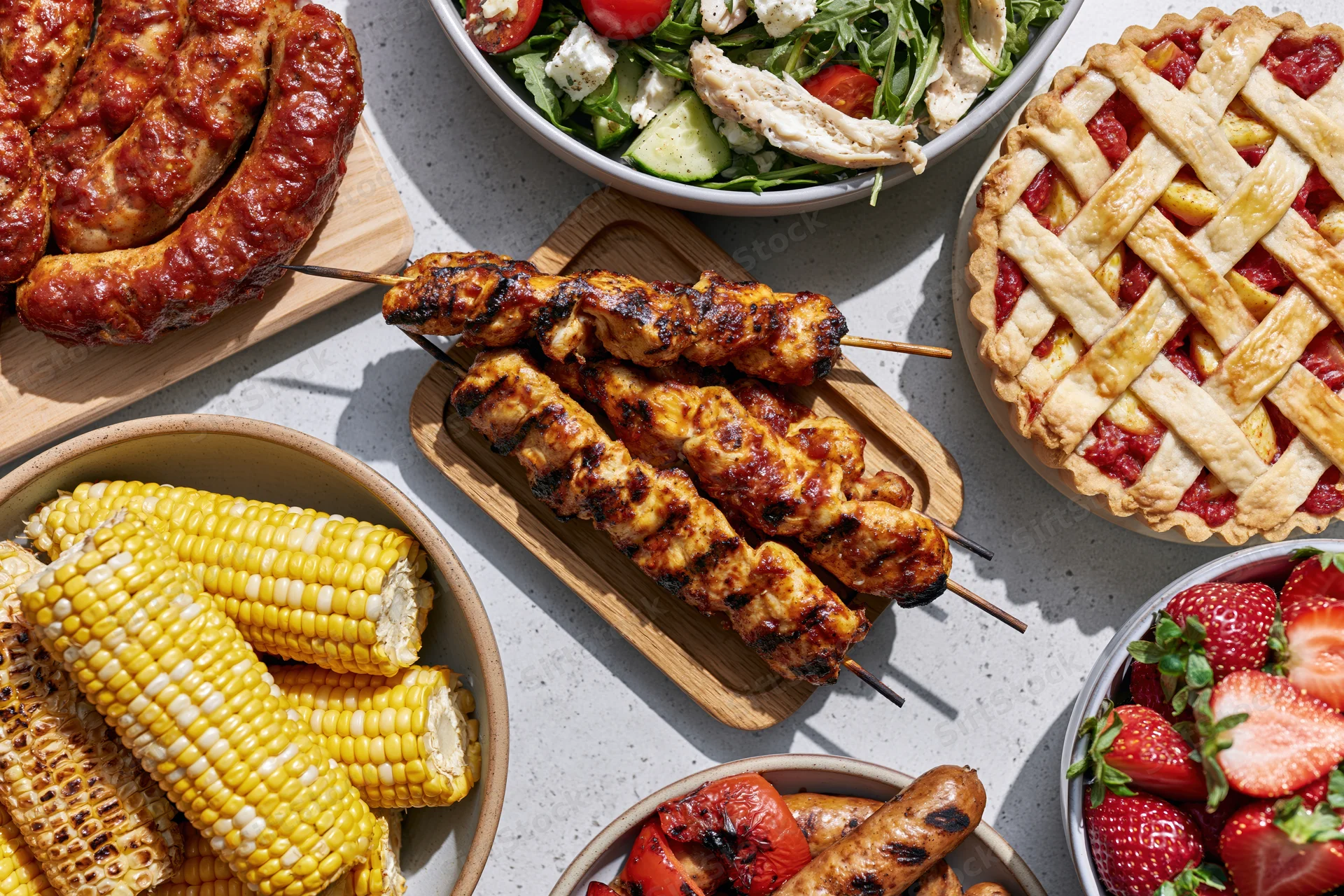 Overhead flat lay of a summer barbecue spread with grilled meats, corn, salad, fruit, and pie on a light gray surface
