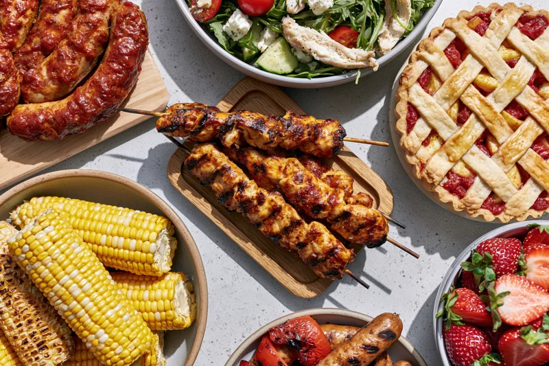 Overhead flat lay of a summer barbecue spread with grilled meats, corn, salad, fruit, and pie on a light gray surface