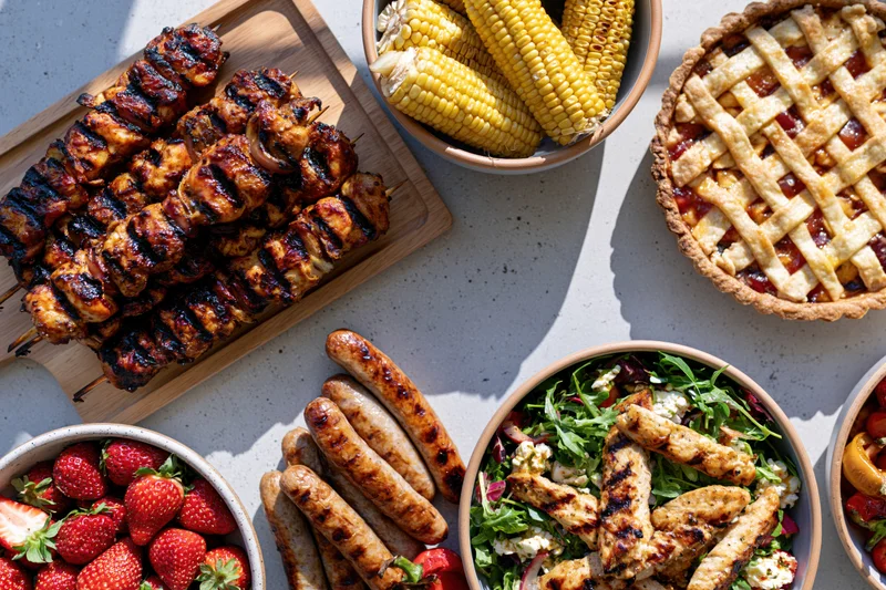 Overhead flat lay of grilled chicken skewers, corn, pie, strawberries, sausages, and salad arranged on a light surface for a summer barbecue
