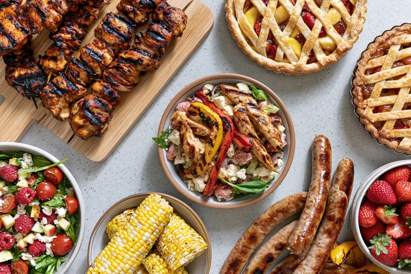 Grilled chicken skewers, sausages, corn, salads and fruit pies arranged on a light countertop for a summer barbecue spread