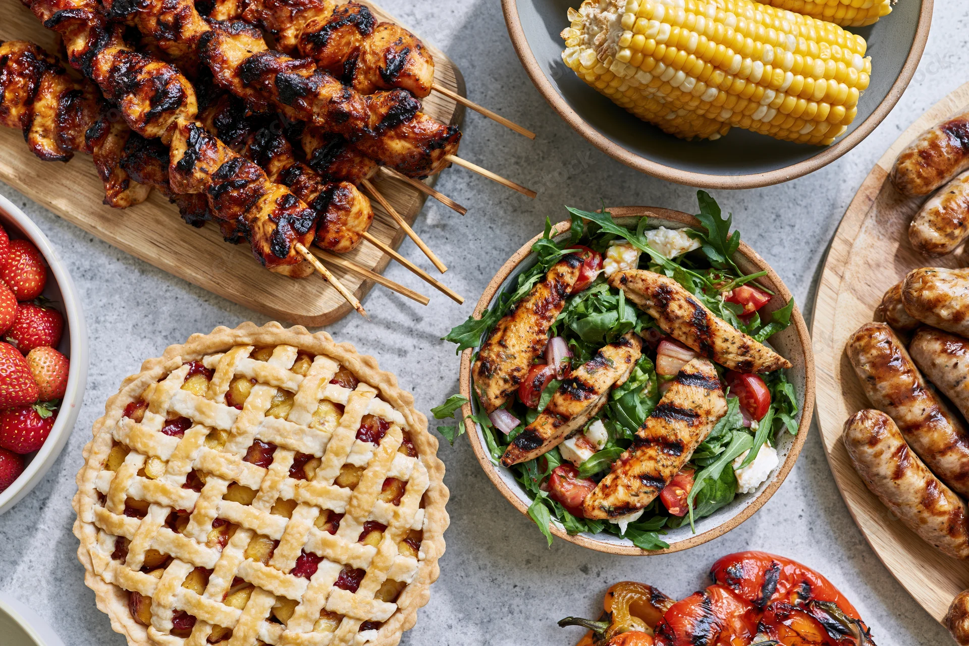 Overhead view of grilled chicken skewers, corn, sausages, fruit, salad, and lattice pie arranged on a gray surface for a summer barbecue spread