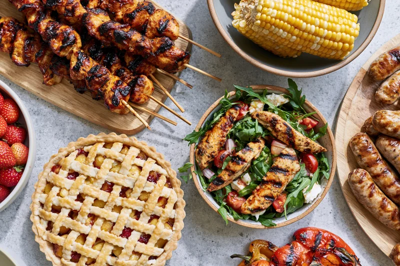 Overhead view of grilled chicken skewers, corn, sausages, fruit, salad, and lattice pie arranged on a gray surface for a summer barbecue spread