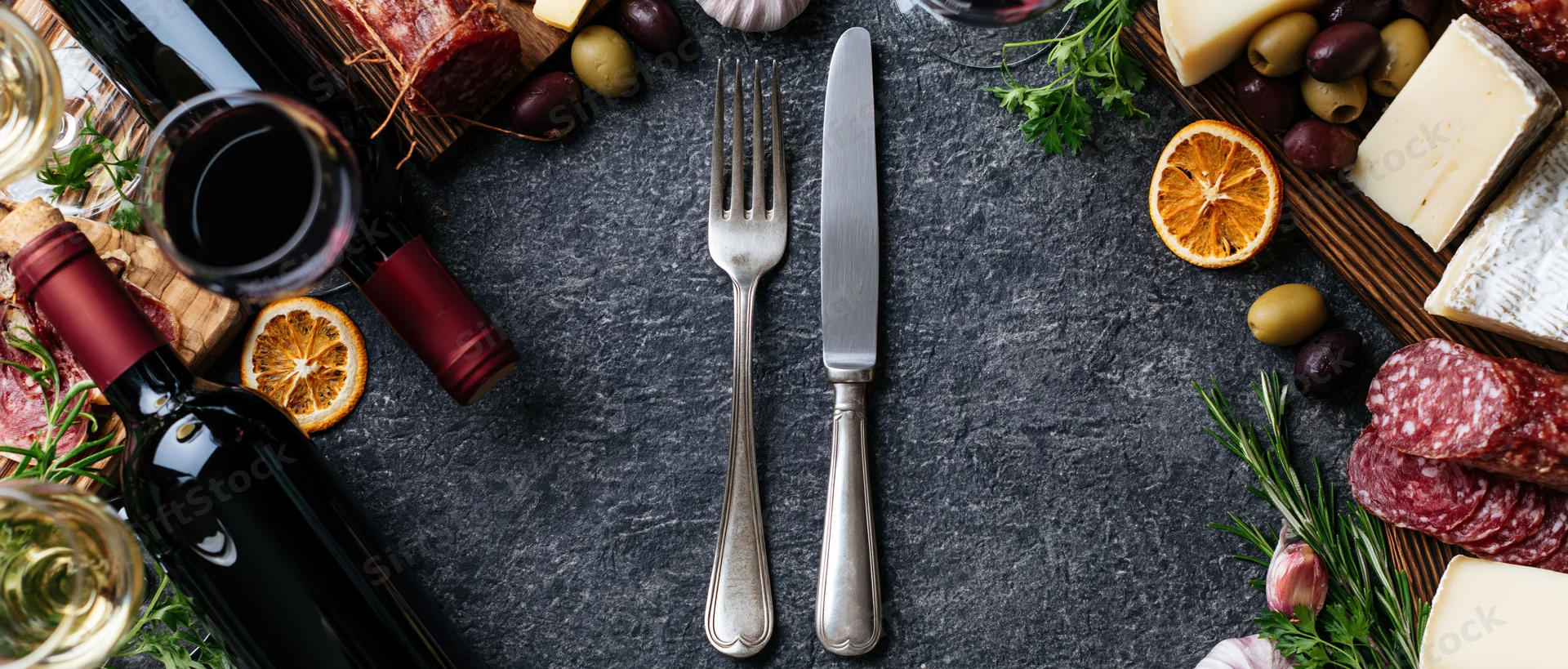 Vintage silver fork and knife centered on dark slate with charcuterie, cheese, wine, and herbs arranged around