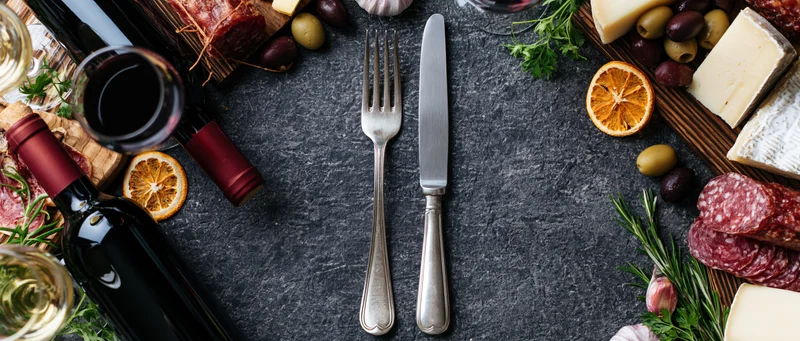 Vintage silver fork and knife centered on dark slate with charcuterie, cheese, wine, and herbs arranged around