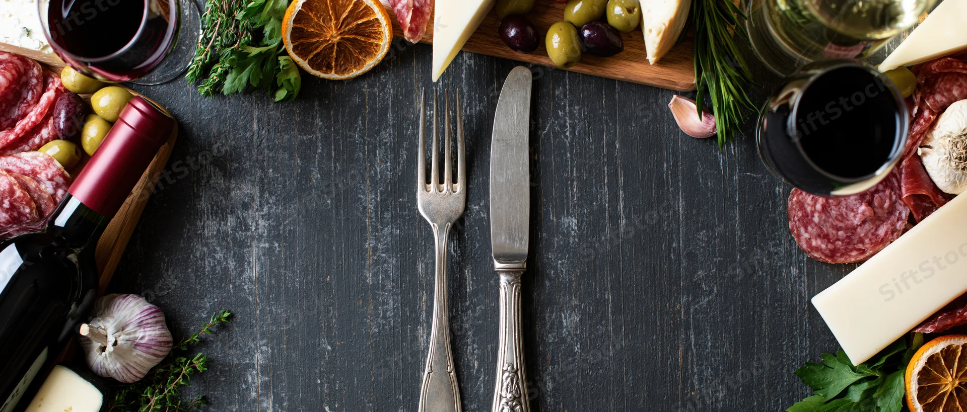 Vintage silver fork and knife centered on dark rustic table surrounded by charcuterie, cheese, wine and herbs