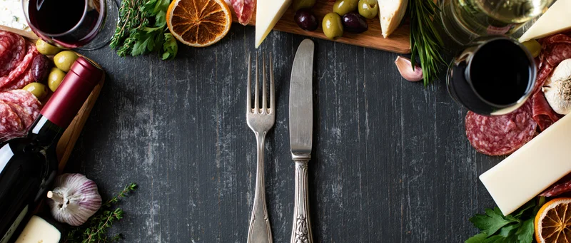 Vintage silver fork and knife centered on dark rustic table surrounded by charcuterie, cheese, wine and herbs