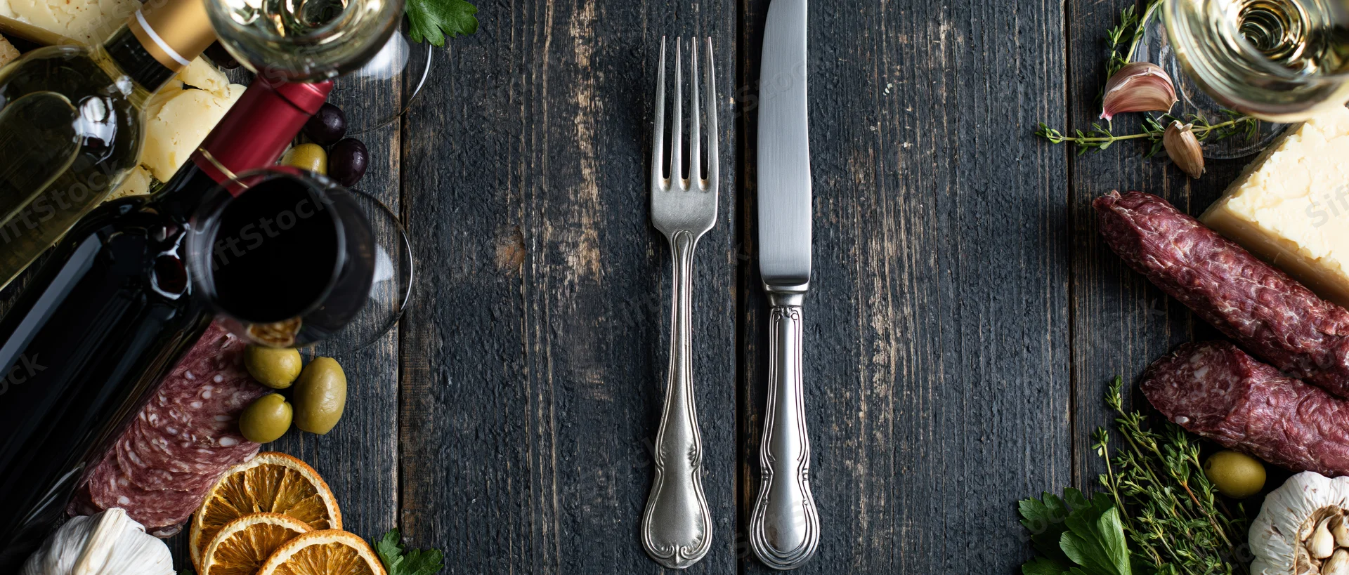 Vintage silver fork and knife centered on dark wooden table surrounded by charcuterie, cheese, olives, wine and herbs