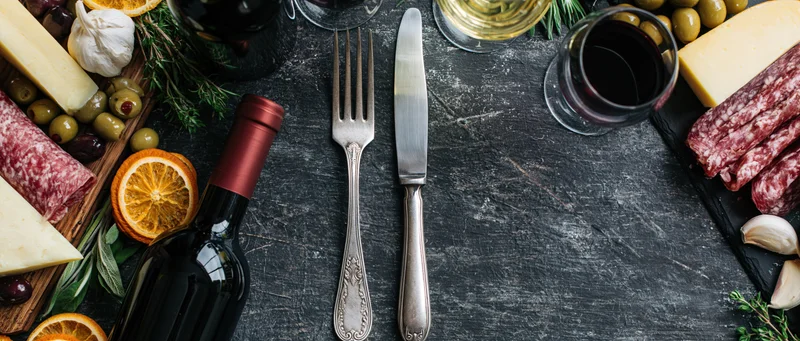 Vintage silver fork and knife centered on dark slate table surrounded by cheese, salami, olives, wine and herbs in overhead flat lay