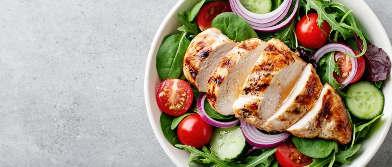 Grilled chicken salad with mixed greens and vegetables in a white bowl on a gray surface