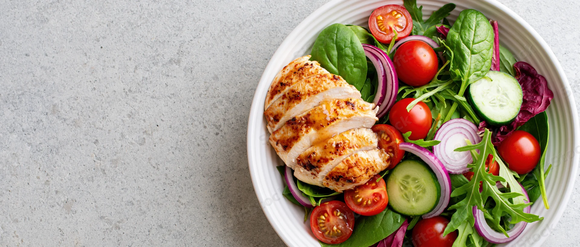 Grilled chicken salad with mixed greens and vegetables in a white bowl on a gray surface