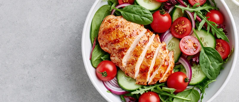 Sliced grilled chicken in a salad bowl with tomatoes cucumbers and onions on a gray surface