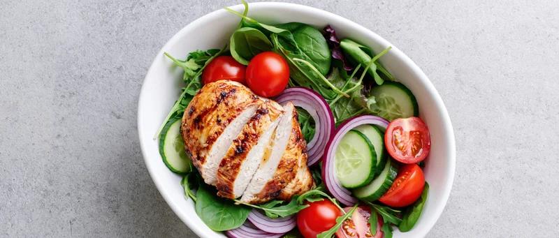 Grilled chicken salad with tomatoes cucumbers and red onions in a white bowl on a gray surface