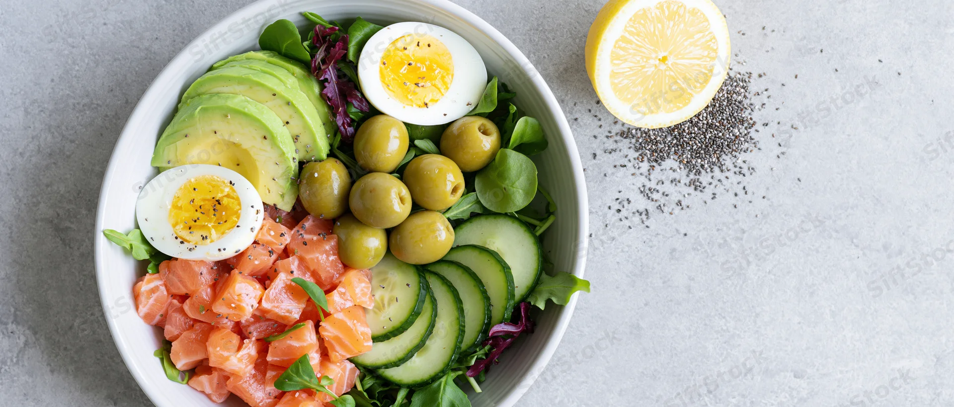 Overhead flat lay of a white ceramic bowl filled with diced salmon, avocado, cucumber, hard-boiled egg, olives and leafy greens on a gray surface with lemon and chia seeds beside it