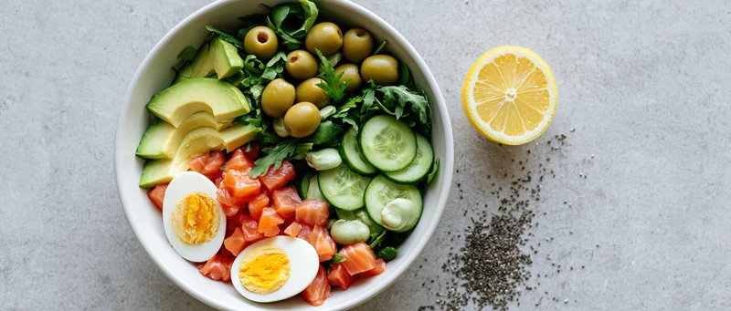 Overhead flat lay of a white ceramic bowl filled with diced salmon, avocado, cucumber, olives, arugula, and hard-boiled eggs, with lemon and chia seeds on the side
