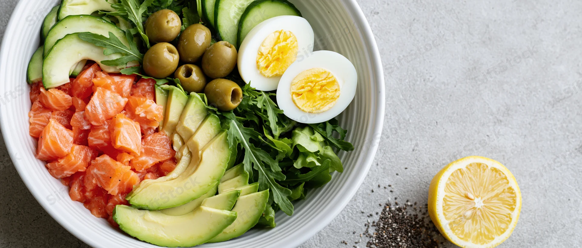 White ceramic bowl filled with salmon, avocado, hard-boiled egg, arugula, cucumber, olives on gray surface with lemon half and chia seeds beside it