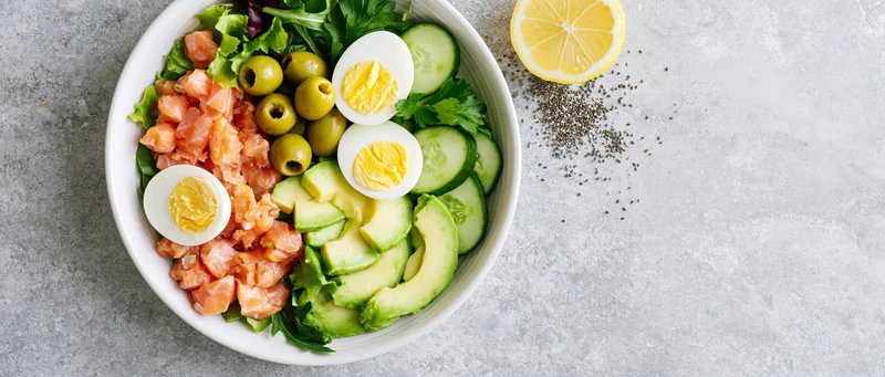 Overhead view of a white ceramic bowl filled with salmon, avocado, hard-boiled eggs, cucumbers, olives and greens on a gray surface with lemon and pepper beside it