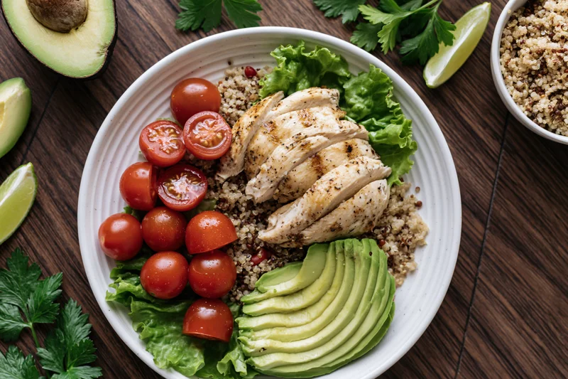 Overhead view of a buddha bowl with quinoa, grilled chicken, avocado, cherry tomatoes, and lettuce on a dark wooden table