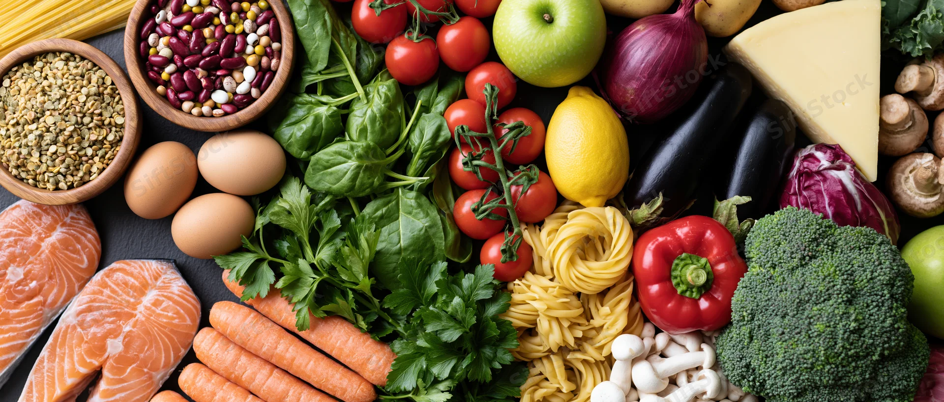 Overhead flat lay of fresh whole foods including vegetables, fruits, protein, and grains arranged on dark surface