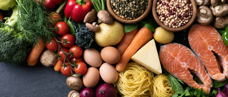 Overhead flat lay of fresh whole foods arranged on dark surface including vegetables, fruits, salmon, eggs, cheese, pasta, mushrooms, and legumes