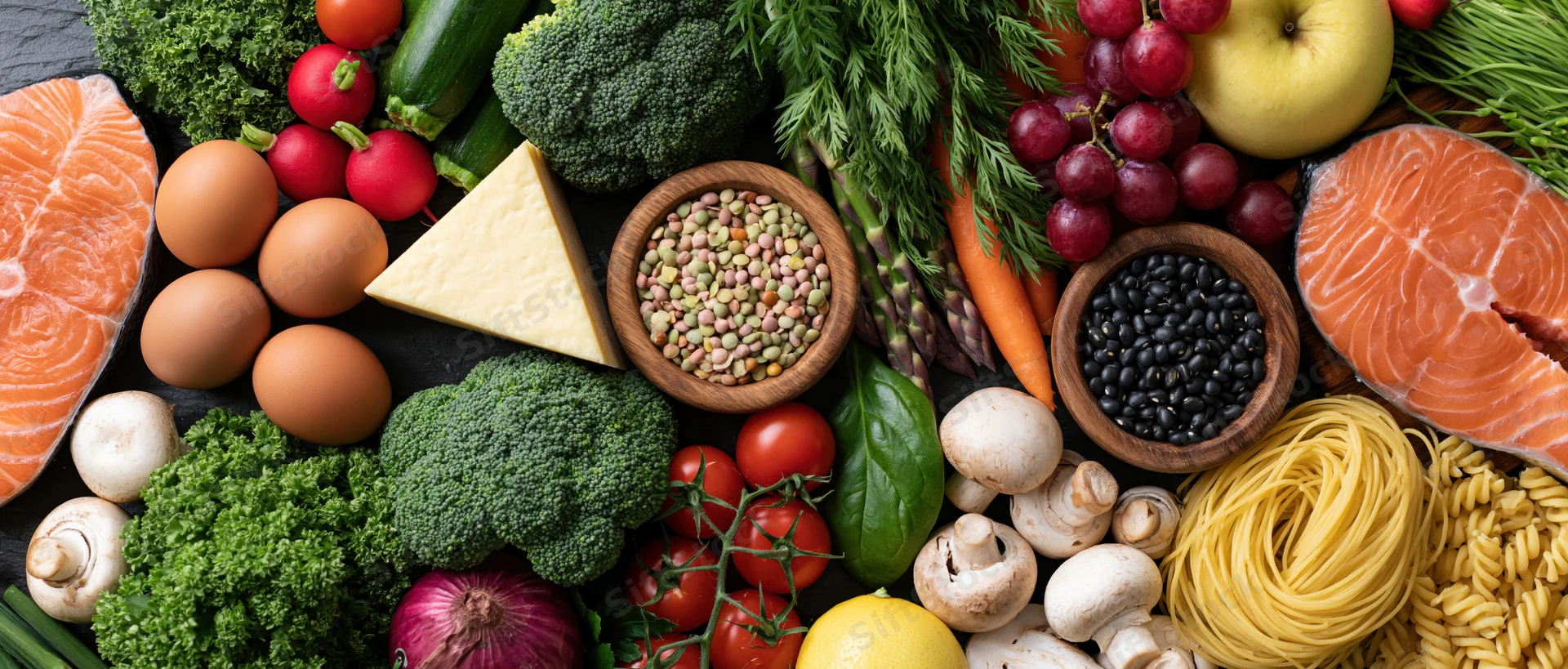 Overhead flat lay of fresh whole foods including salmon, eggs, vegetables, fruits, grains, and dairy arranged on dark surface