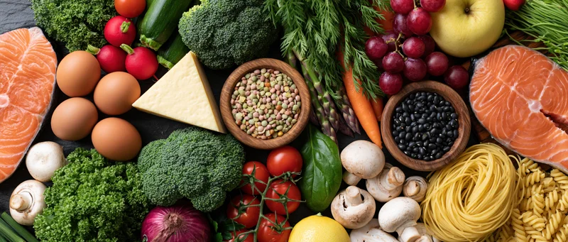 Overhead flat lay of fresh whole foods including salmon, eggs, vegetables, fruits, grains, and dairy arranged on dark surface