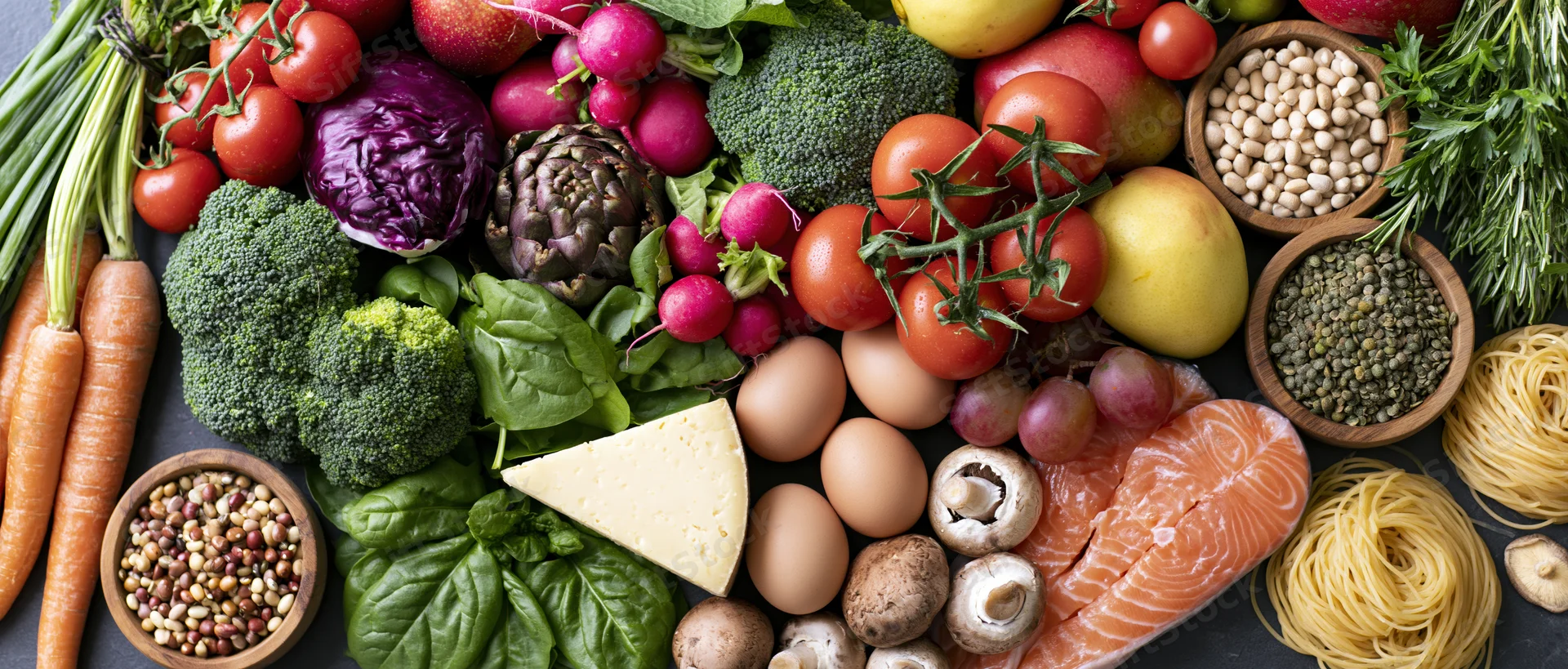 Overhead flat lay of fresh whole foods including vegetables, fruits, protein, and grains arranged on dark surface