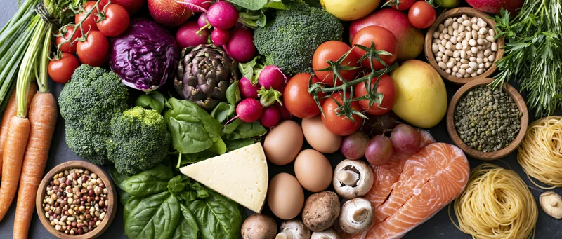 Overhead flat lay of fresh whole foods including vegetables, fruits, protein, and grains arranged on dark surface