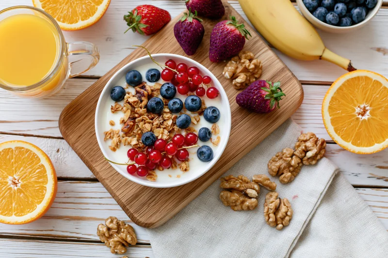 Yogurt bowl topped with granola, blueberries, strawberries, redcurrants, and walnuts on a wooden cutting board with orange slices, banana, and juice on a white wooden table
