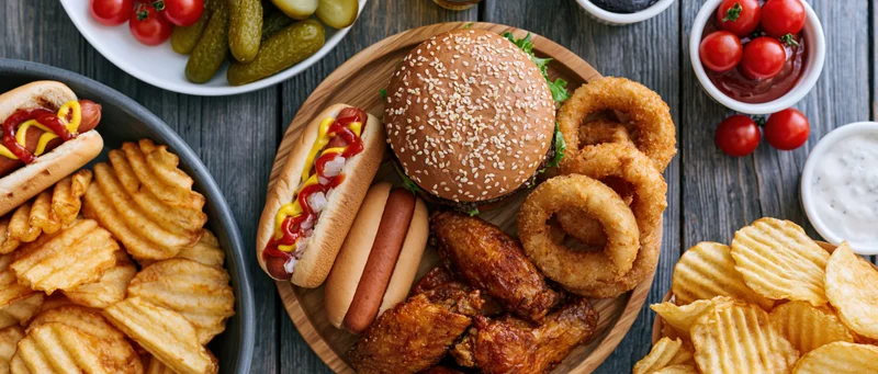 Overhead flat lay of american casual dining spread with burger, hotdogs, fries, onion rings and chicken wings on dark wooden table