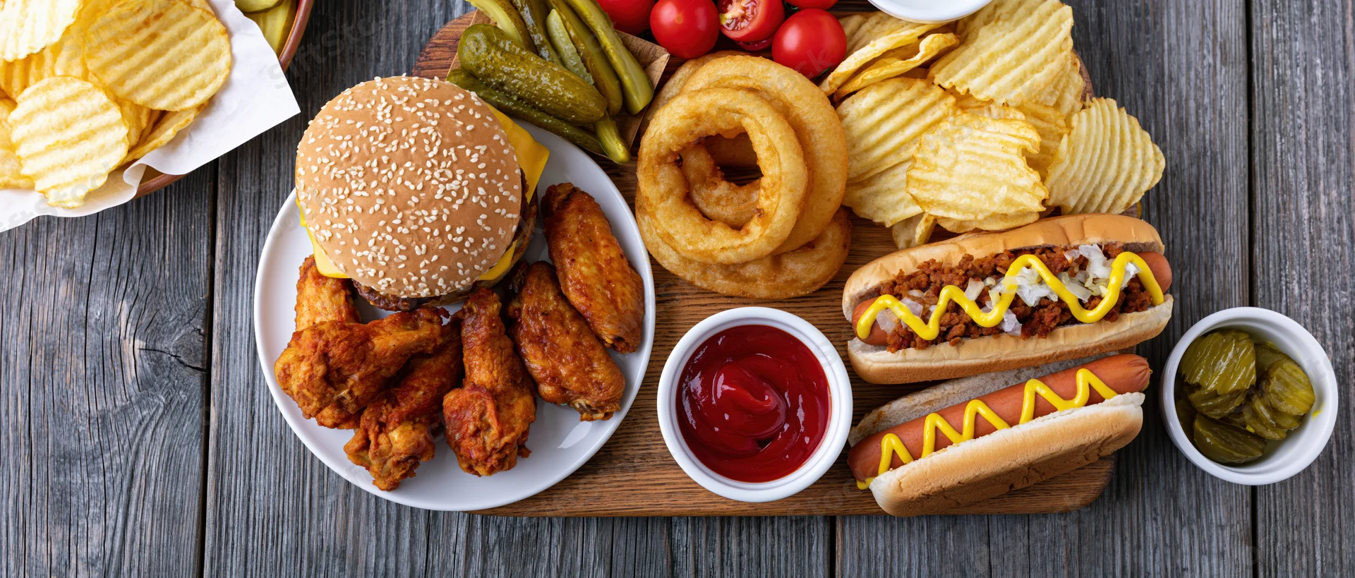 Overhead flat lay of american casual dining spread with burger, hotdogs, fries, onion rings, chicken wings, and condiments on wooden table