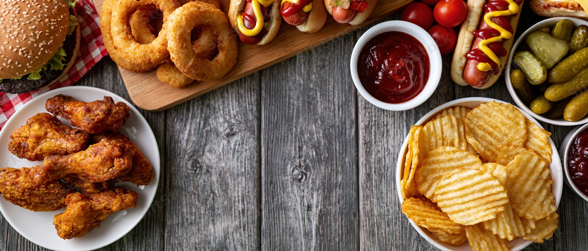 Overhead flat lay of american fast food spread on wooden table with burger, hotdogs, fries, onion rings, chicken wings, chips, pickles, ketchup and mustard
