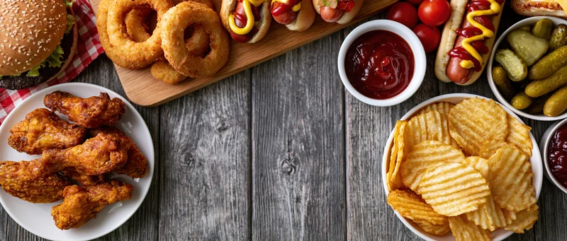 Overhead flat lay of american fast food spread on wooden table with burger, hotdogs, fries, onion rings, chicken wings, chips, pickles, ketchup and mustard