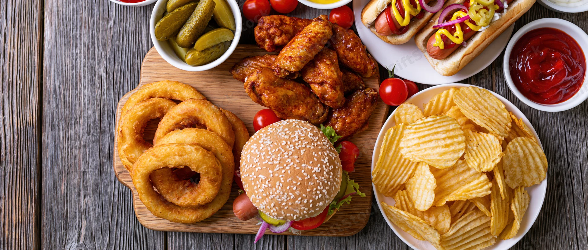 Overhead flat lay of american casual dining spread with burger, fries, onion rings, chicken wings, hot dogs, and condiments on wooden table