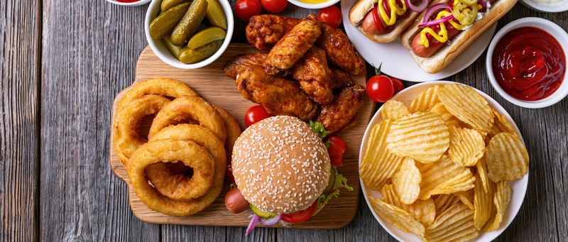 Overhead flat lay of american casual dining spread with burger, fries, onion rings, chicken wings, hot dogs, and condiments on wooden table
